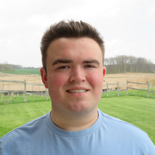 Andrew is wearing a blue t-shirt. He is standing in a front of a wooden fence in an open field of grass. He is smiling at the camera. 