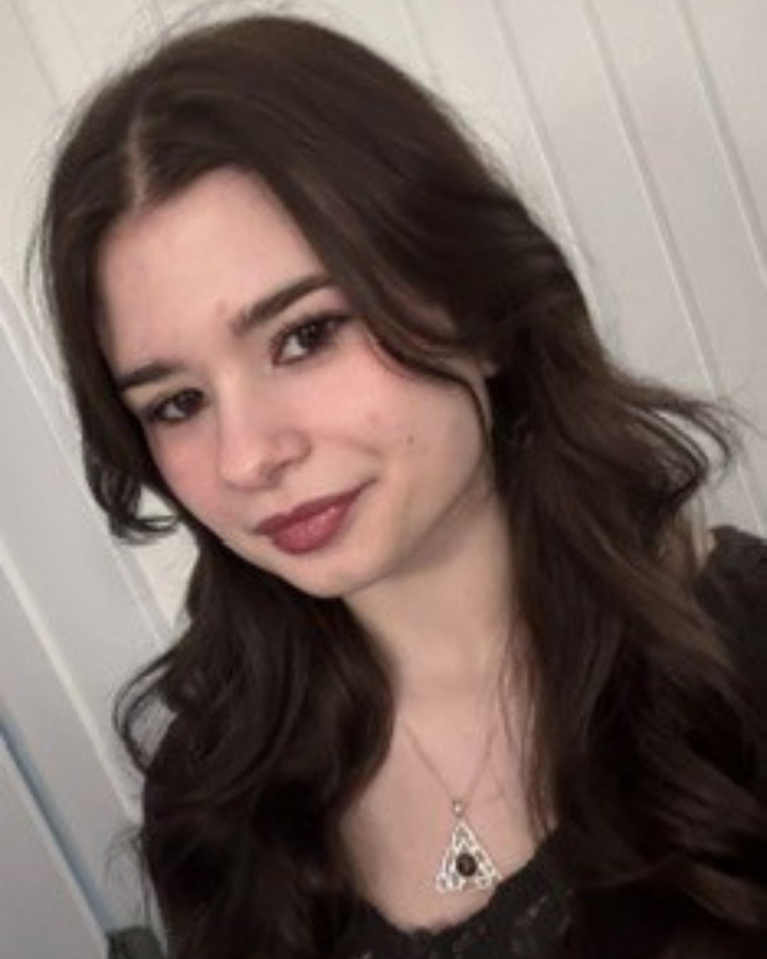 Catherine is smiling for a selfie that shows her from the shoulders up. She has long, dark hair and is wearing a dark shirt with a silver necklace.