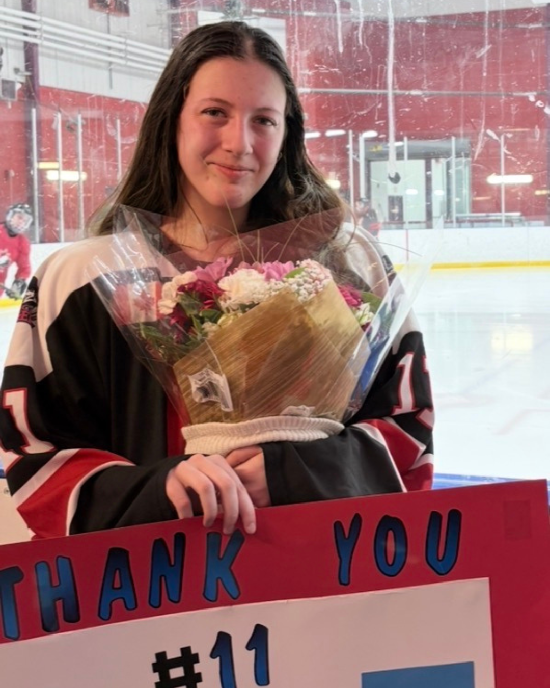 Chelsea is posing for a photo after playing in a hockey game. She is still wearing her gear as she sands beside the glass holding flowers and a Thank You #11 sign.