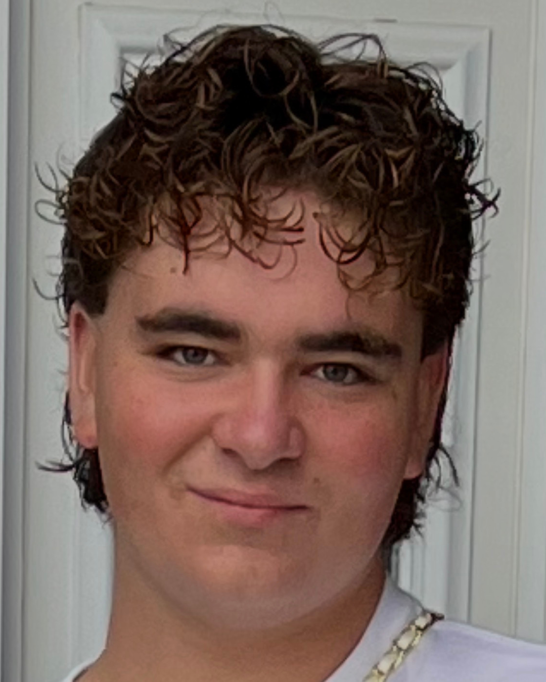 Chipp is smiling for a selfie that shows him from the shoulders up, He has brown messy hair and is wearing a white shirt and gold chain.
