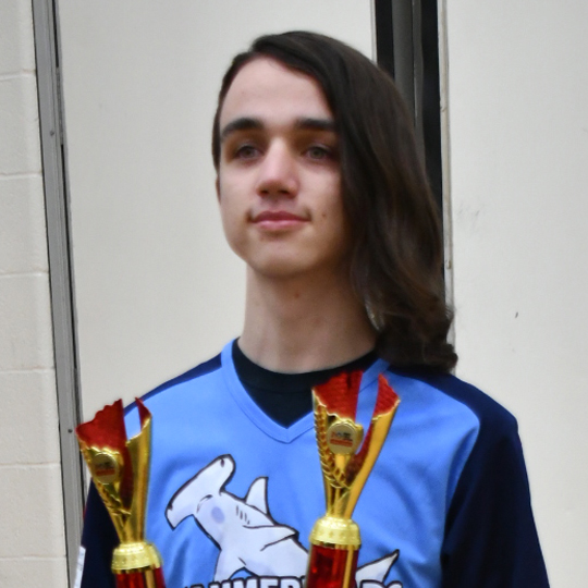 Corben is wearing a blue Hammerheads jersey with dark pants and black sneakers. He is standing and posing with two golden and red trophies. He is in a gymnasium with light brown flooring and a white wall behind him. 