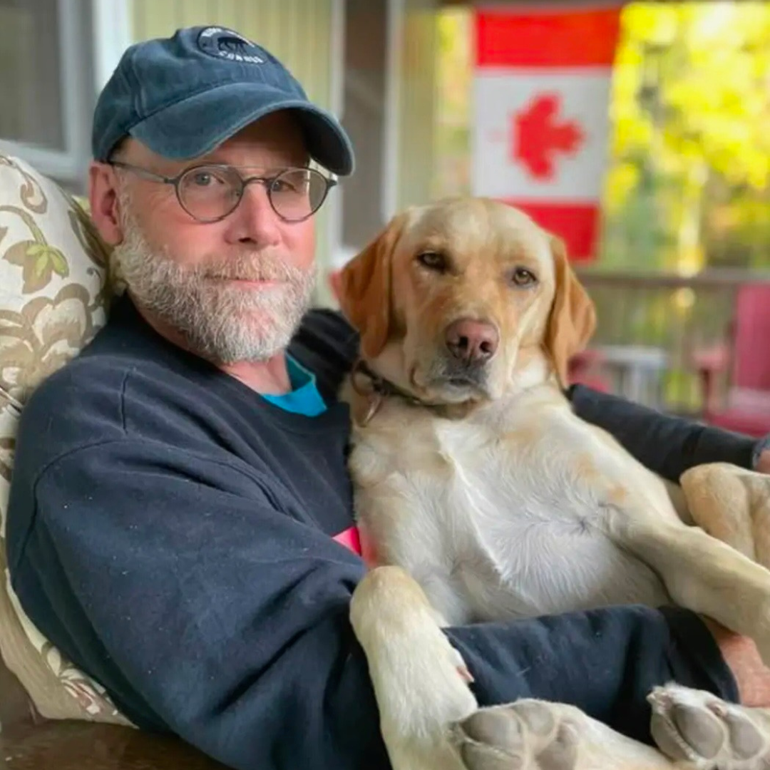 David Schaller is wearing a dark sweater, a blue baseball hat and dark glasses in this photo. He is sitting on a brown chair on a deck and a Canadian flag can be seen in the background.