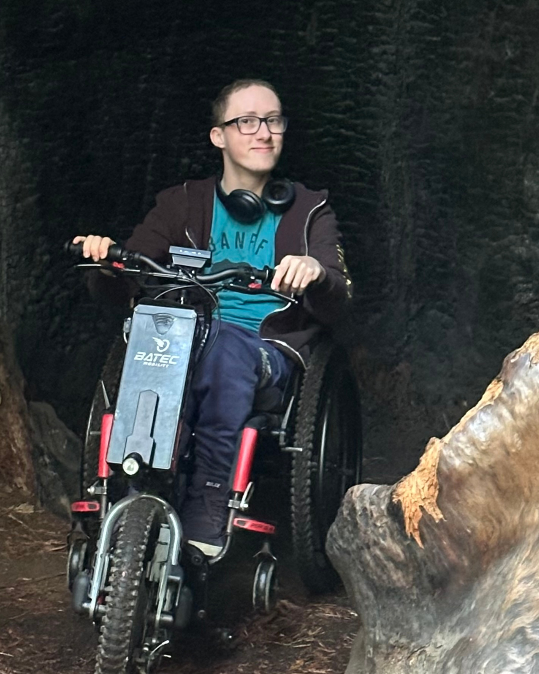 Hayden is shown in a large redwoods forest. His wheelchair is adapted with a motorbike attachment to navigate through the woods.