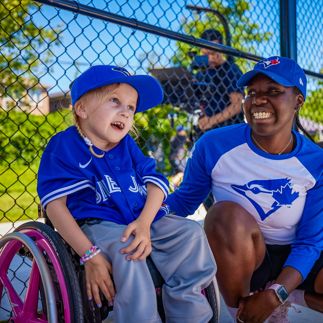 A young girl is smiling for a photo while sitting in her pink wheelchair. An adult woman is crouched next to her and smiling at the young girl. 