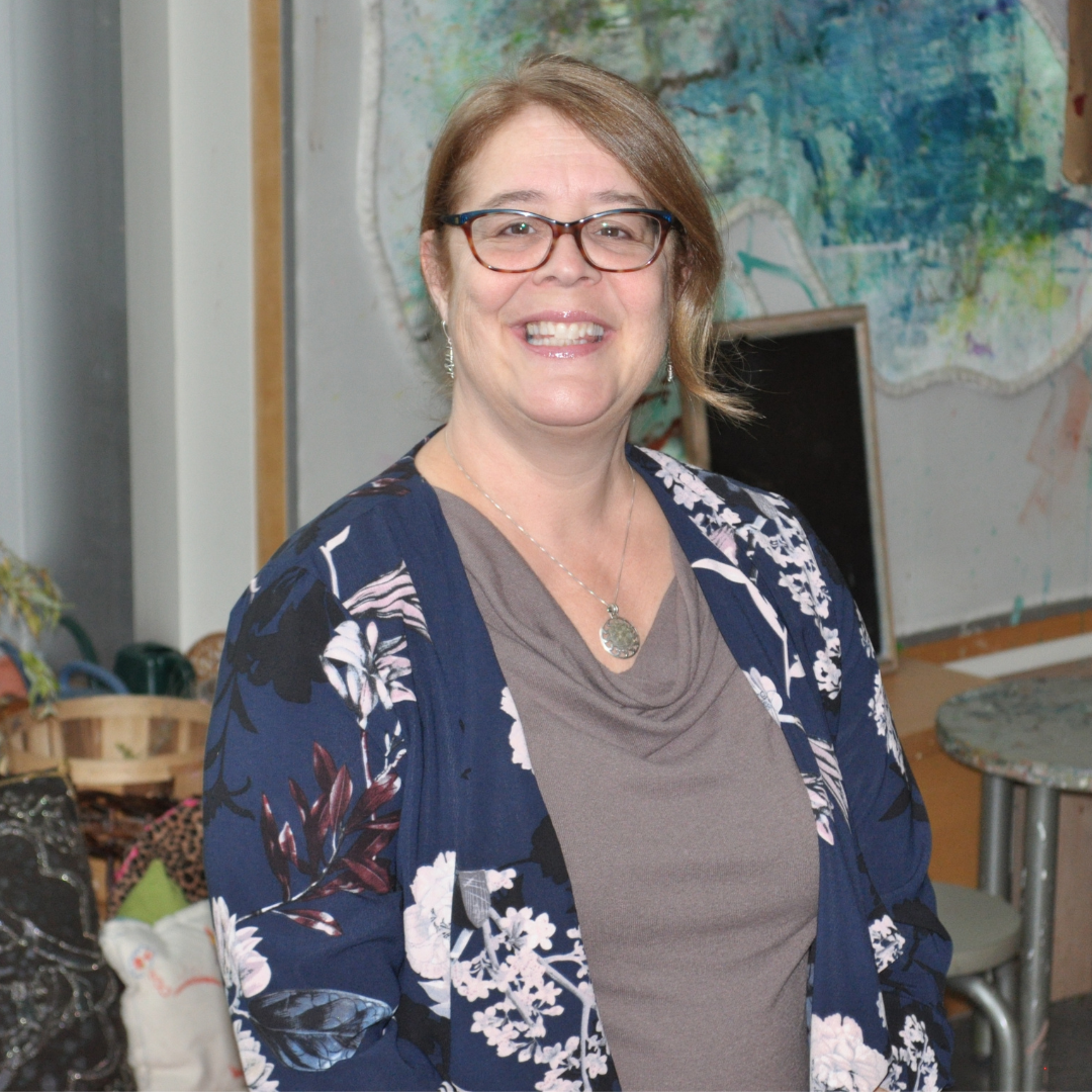 Jessica is smiling at the camera. She is wearing a dark blue cardigan with a floral design and a grey shirt with a scooped neckline. She has on a silver necklace with a pendant and is wearing dark glasses. In the background there is artwork and stools.