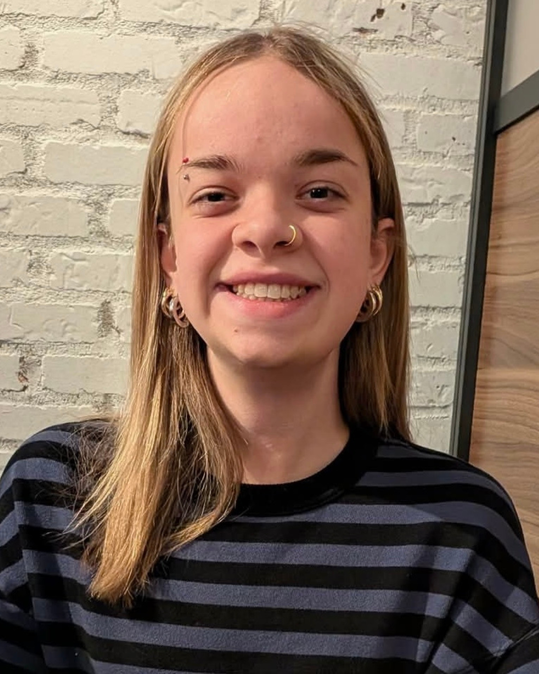 Jocelyne is smiling for a photo in front of a painted white wall. She has long, blond hair and an eyebrow, nose and multiple ear piercings. She is wearing a blue and black striped shirt.