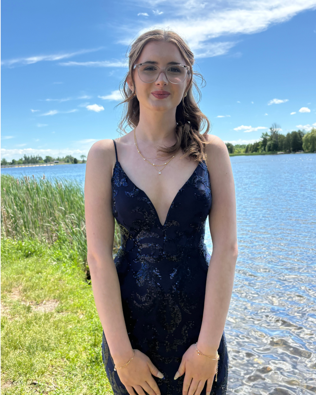 Kara is posing for a photo in her prom dress. The dress is long and dark blue, and her brown hair is styled in waves. She is standing in front of a beautiful lake scene.