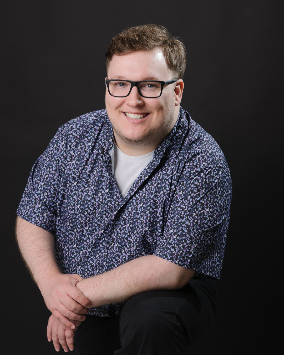 Liam is smiling for a professional photo. He is leaning forward on his left knee and wearing a patterned blue shirt and dark pants. The background is a dark grey.