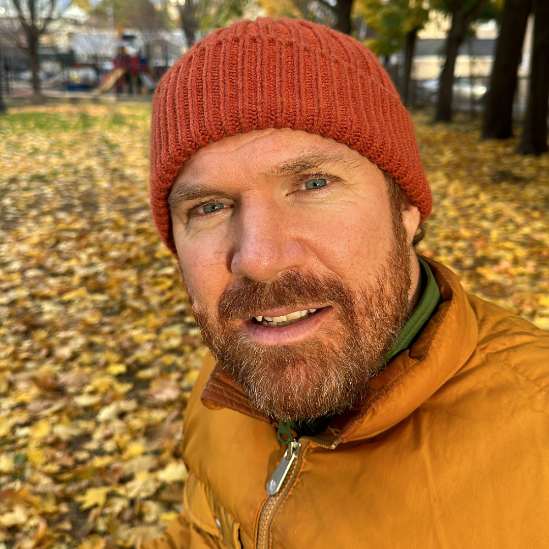 Luke is wearing a red beanie and a beige jacket. He is sitting in a park with leaves covering the ground and trees in the background.
