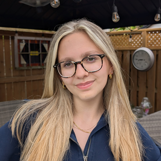 Rebecca is wearing a dark blue hoodie and has on dark glasses and golden hoop earrings. She is smiling at the camera and standing in front of a backyard patio with dangling lights above her head. 