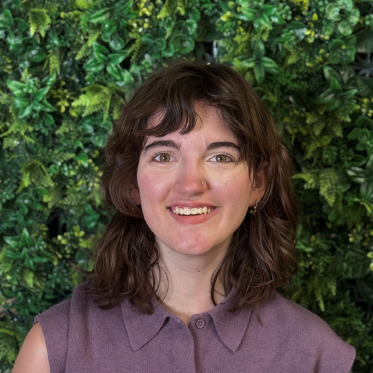 Sable is wearing a short-sleeved purple button down with silver earrings and a nose stud. She is smiling into the camera and is standing in front of a faux green bush background. 