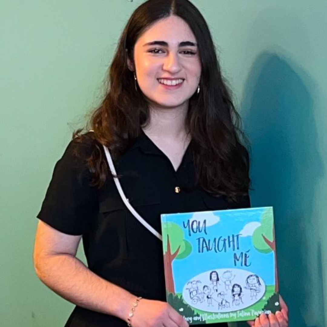 Talina is holding her book, You Taught Me, in this picture. She is wearing a black blouse with see-through, striped sleeves. She is standing in front of a concrete wall