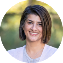 Anchel is smiling for her headshot with a summer day blurred in the background. Her hair is short and brown, and she is wearing a light coloured shirt under a white cardigan.
