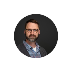 Brendan Wylie-Toal looks pleasantly at the camera in front of a dark grey background. He uses glasses and is wearing a blue plaid shirt under a brown cardigan.