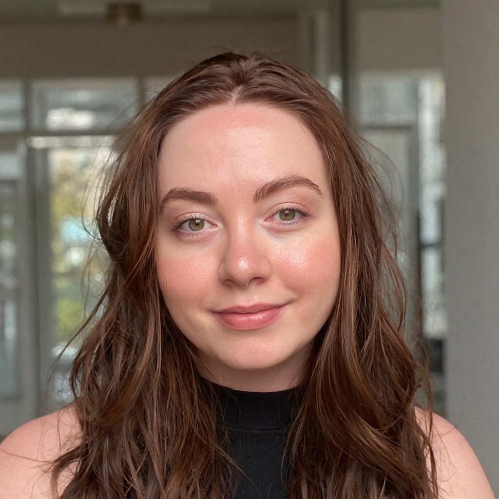 June is smiling at the camera for her headshot. Her hair is brown and wavy, she is wearing a black sleeveless top and the background is blurred.