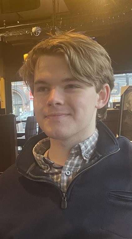 Declan is wearing a dark blue half-zip sweater with a grey flannel button down underneath. He is looking to the side and smiling. He is sitting in a venue with a window behind him. 