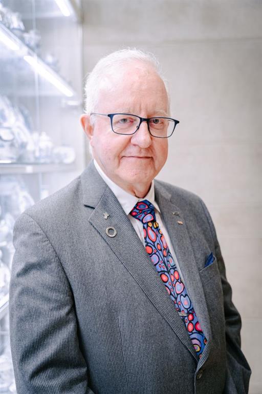 Dr. Peter Rumney is posing for a photo wearing his EKO Tribute Award pinned on his breast pocket. He is wearing a grey jacket with a white collared shirt and an Indigenous art tie.