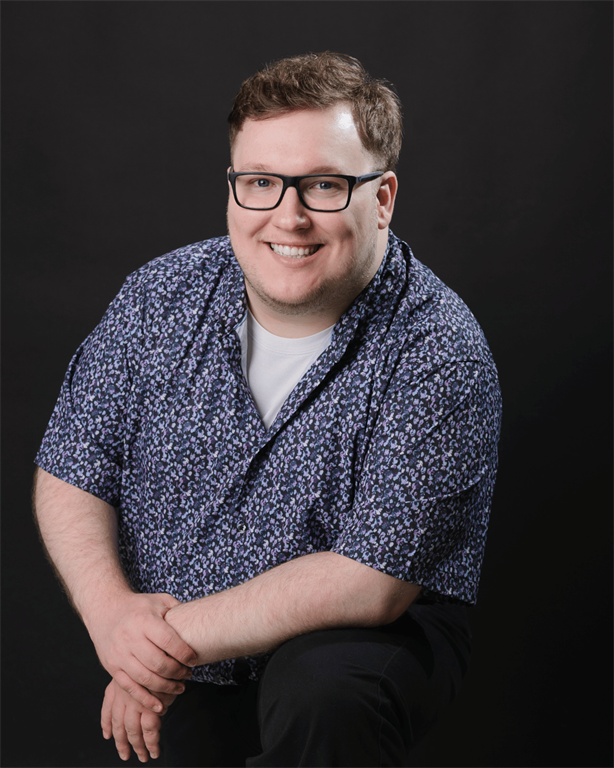 Liam Pickells is leaning forward with his elbows on his left knee as he smiles for a photo in front of adark background.