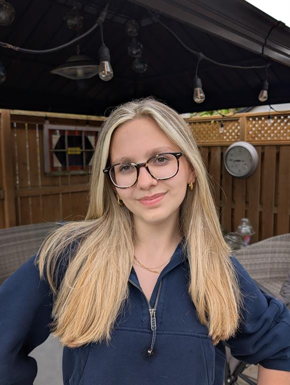 Rebecca is wearing a dark blue hoodie and has on dark glasses and golden hoop earrings. She is smiling at the camera and standing in front of a backyard patio with dangling lights above her head. 