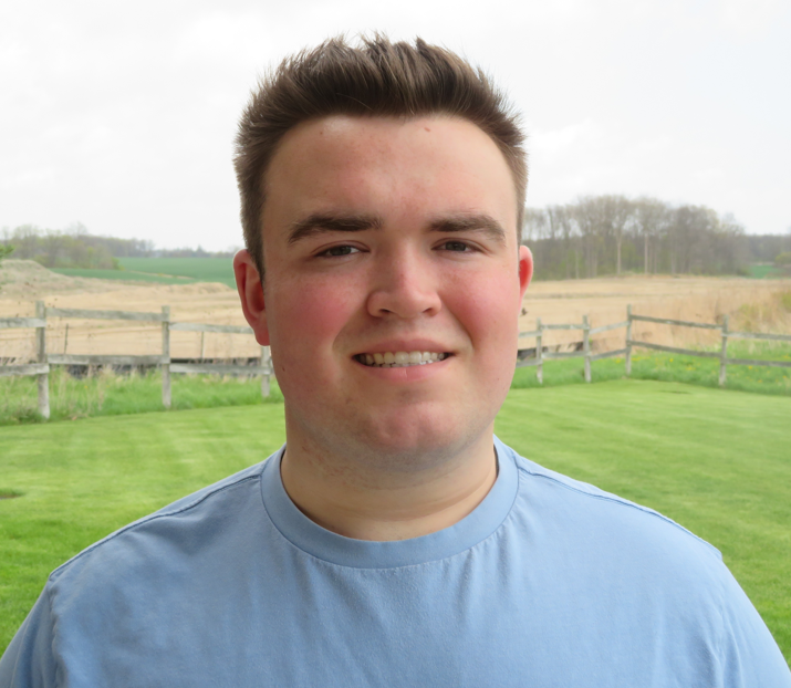 Andrew is wearing a blue t-shirt. He is standing in a front of a wooden fence in an open field of grass. He is smiling at the camera. 