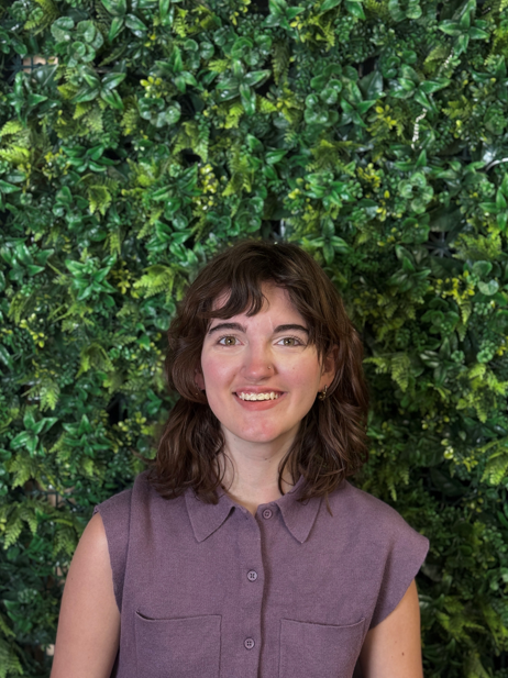 Sable is wearing a short-sleeved purple button down with silver earrings and a nose stud. She is smiling into the camera and is standing in front of a faux green bush background. 