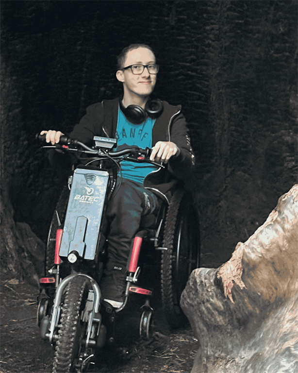 Hayden Tange is sitting in his wheelchair that has been adapted with a motorbike attachment. He is posing inside a large redwood forest.